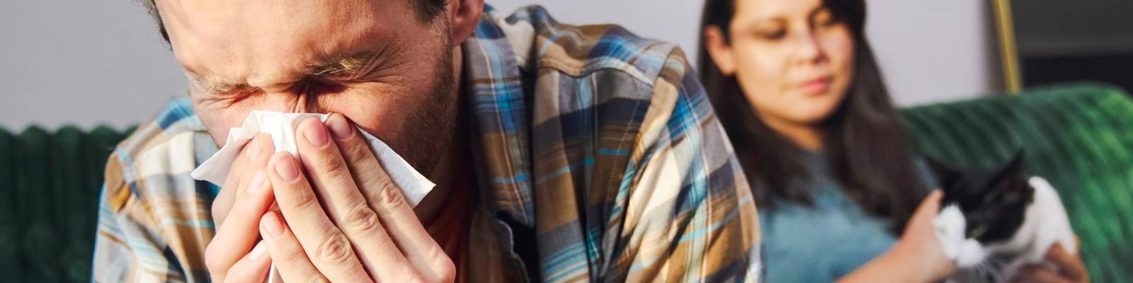 Man suffering from pet allergies sneezes into a tissue while sitting next to a woman holding a cat.