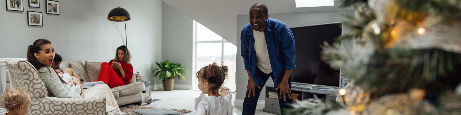 A young African American father is playing with his two young daughters by their Christmas tree in a white colored living room.